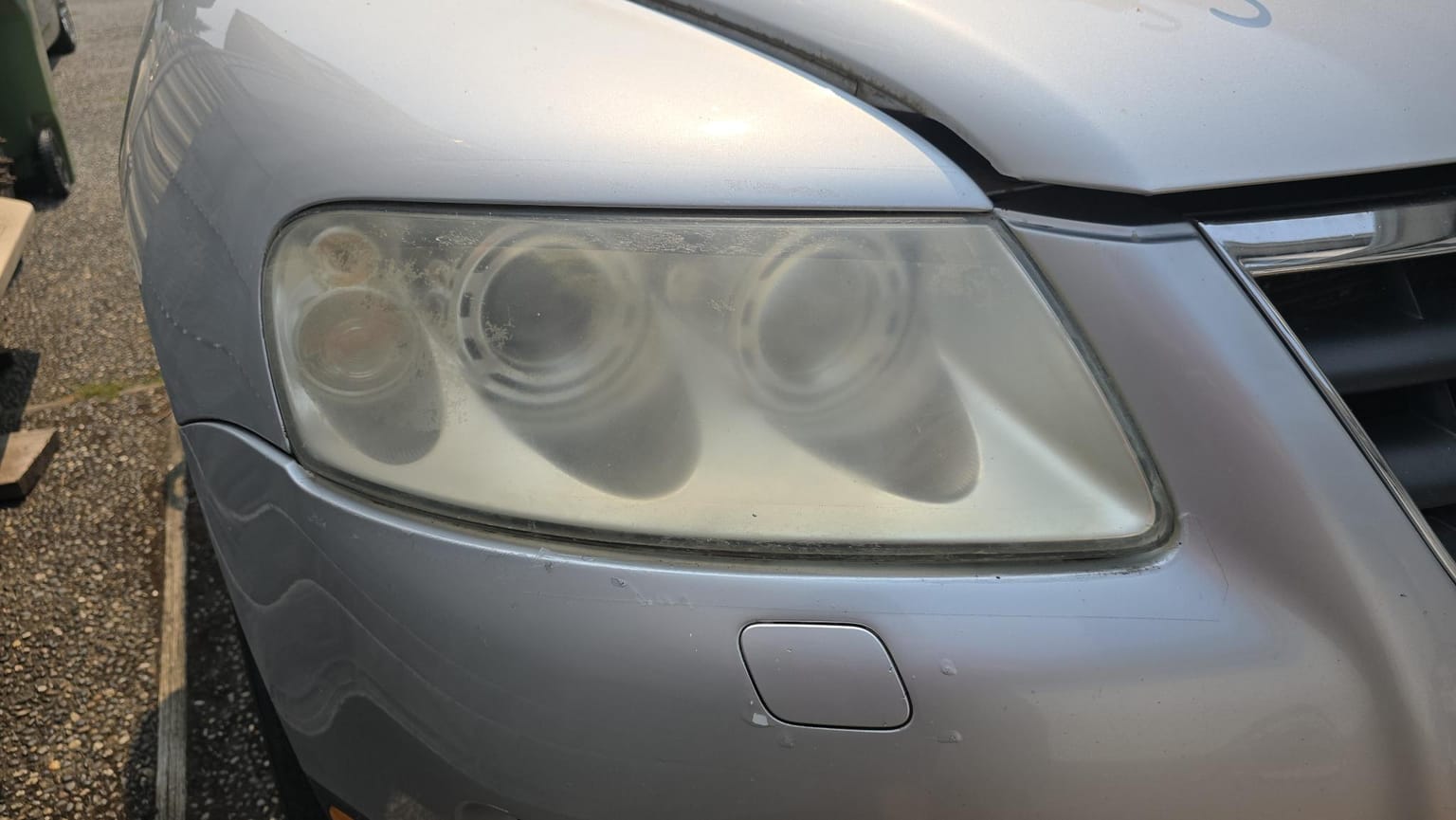 A headlight on a silver sedan before restoration: the lens is yellowed and clouded, and the reflectors behind it are barely visible.